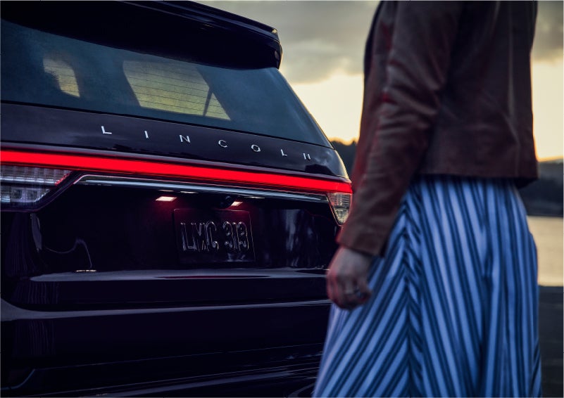 A person is shown near the rear of a 2023 Lincoln Aviator® SUV as the Lincoln Embrace illuminates the rear lights | Duncan Lincoln in Blacksburg VA